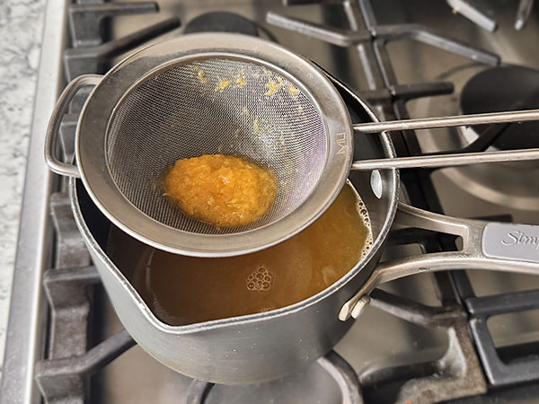 A small saucepan with orange juice and fine mesh strainer on top with some pulp.