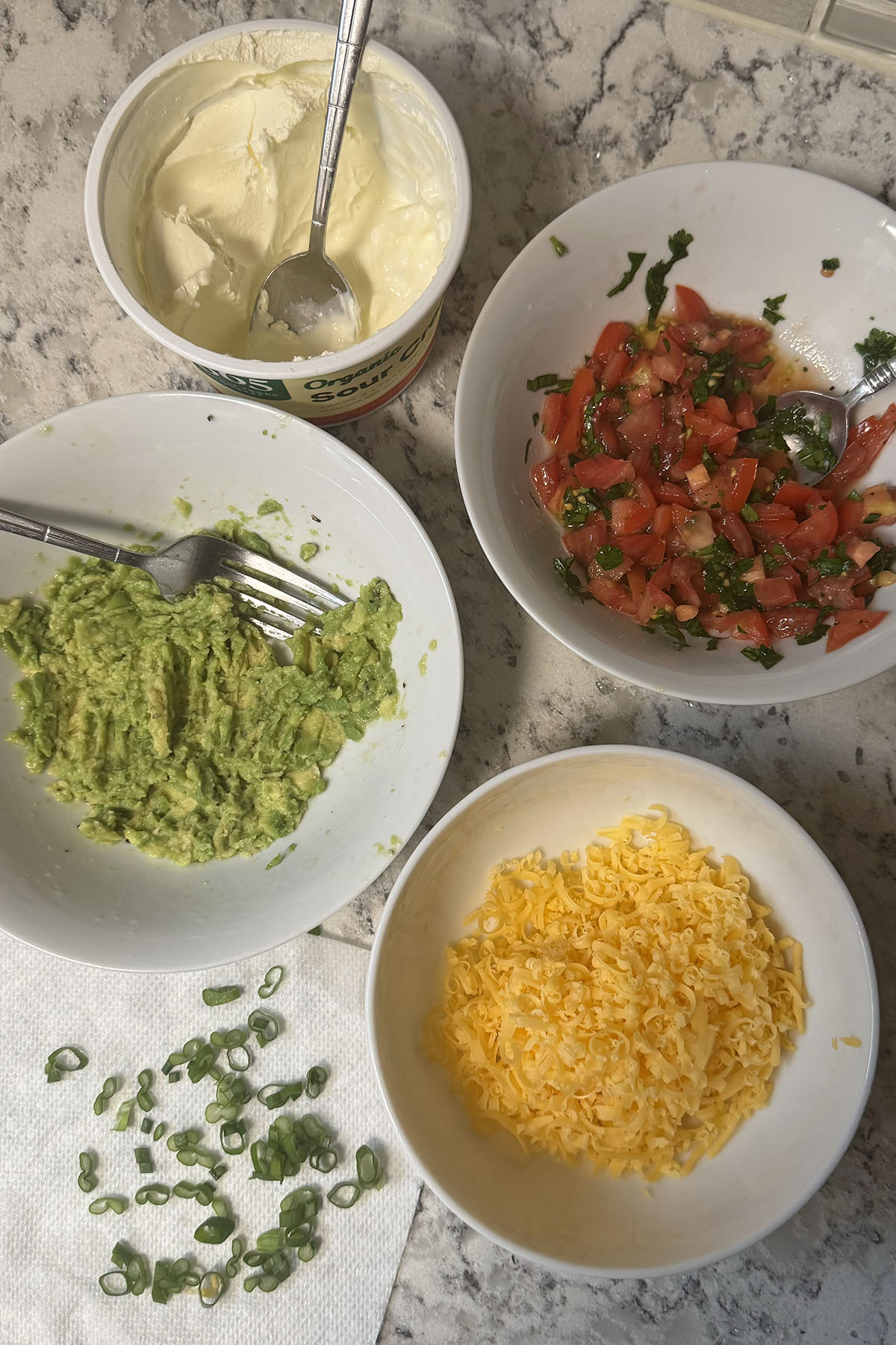 Prepared potato toppings in small bowls: mashed avocado, tomato salsa, grated cheddar, sliced green onions and sour cream.