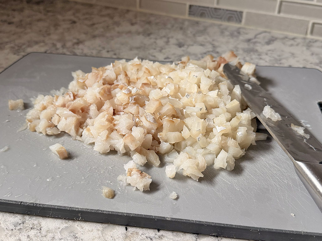 Diced fresh cod fillets on a cutting board with a knife, showing small hand-cut pieces of cod prepared for fish cakes.