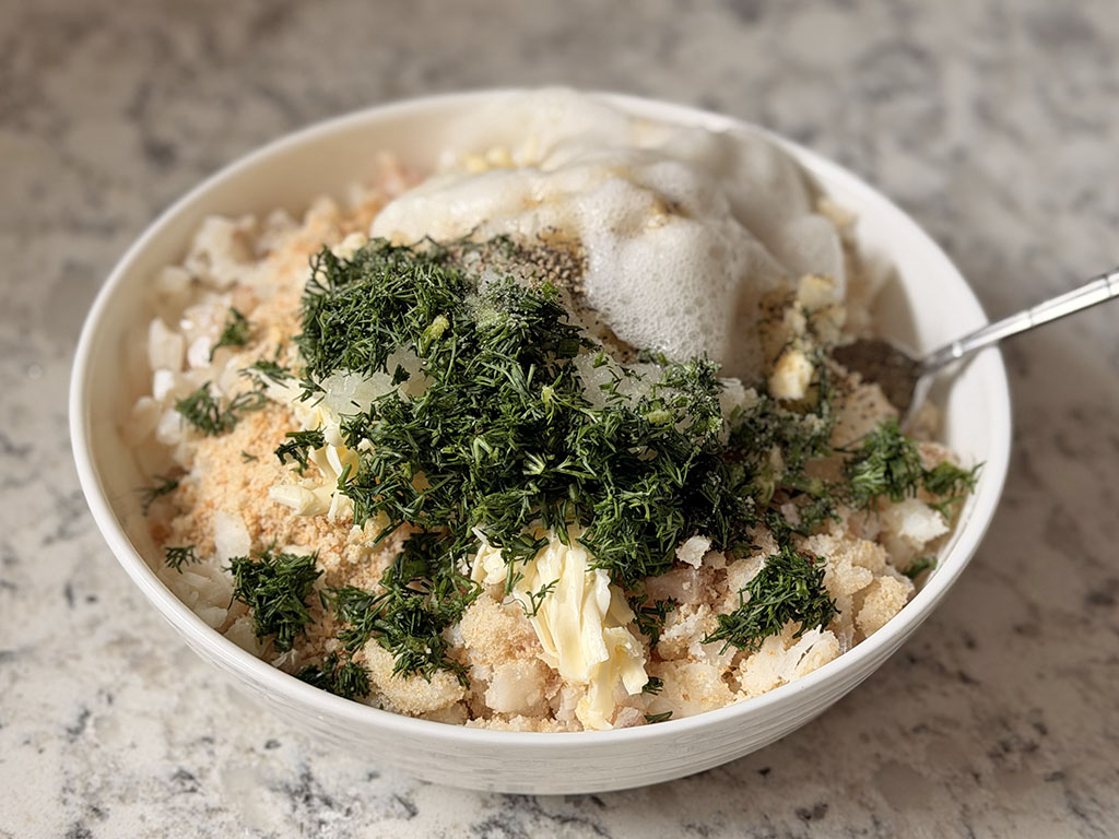 Diced cod in a large bowl with grated onion, grated butter, breadcrumbs, fresh dill, spices, and egg whites added before mixing.