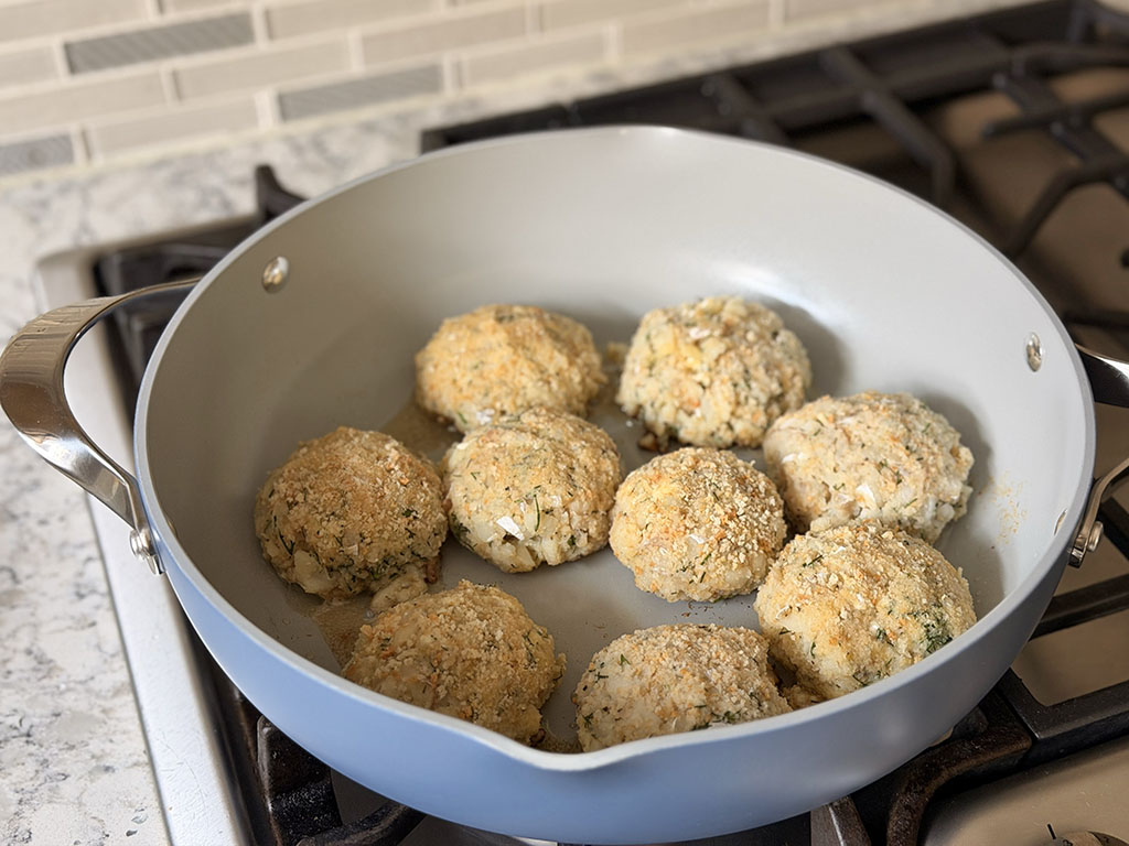 Cod fish cakes cooking in a skillet, browning on the first side while frying over medium heat.