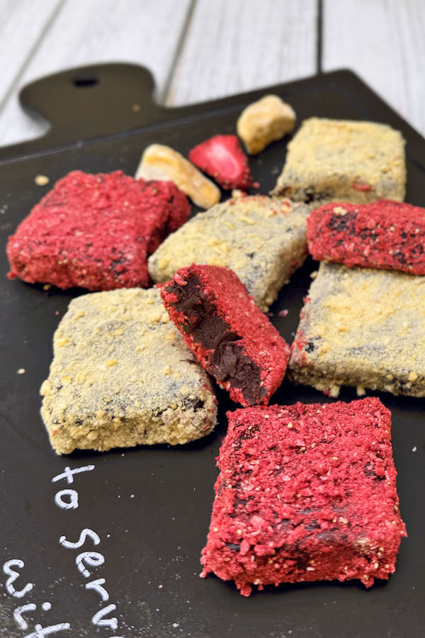 Chocolate truffles rolled in mango and strawberry dust on a serving board, one truffle has a bite taken out of it.