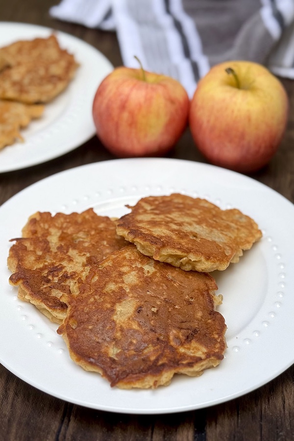 A serving of apple cinnamon pancakes on a plate with fresh apples and another plate of pancakes in the background.