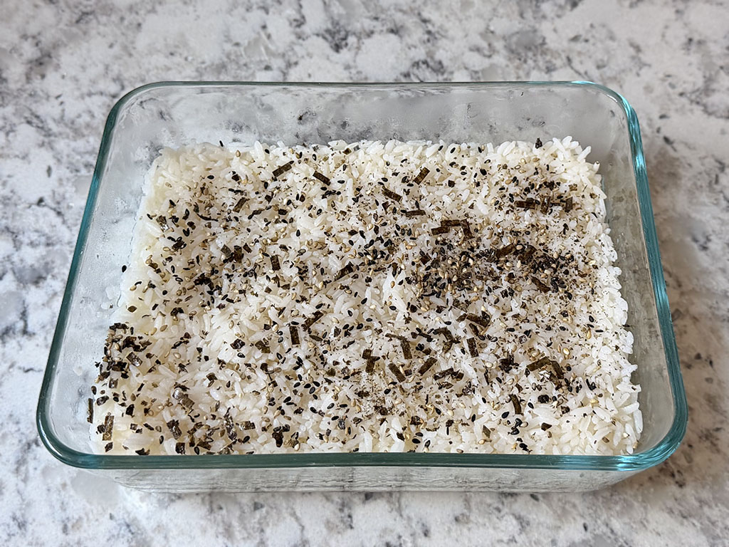 Seasoned rice mixed with rice vinegar, maple syrup, and salt spread evenly in a glass baking dish and topped with furikake seasoning.