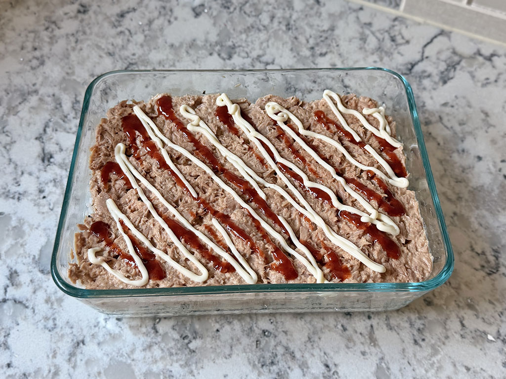 Tuna mixture spread evenly over the rice layer and drizzled with thin lines of mayonnaise and sriracha sauce.