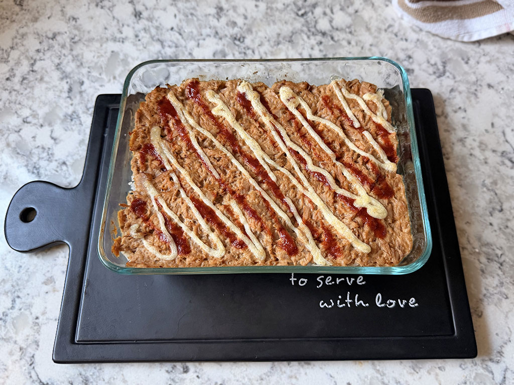 Canned tuna sushi bake fresh from the oven, showing a warm golden tuna layer drizzled with mayonnaise and sriracha over seasoned rice.