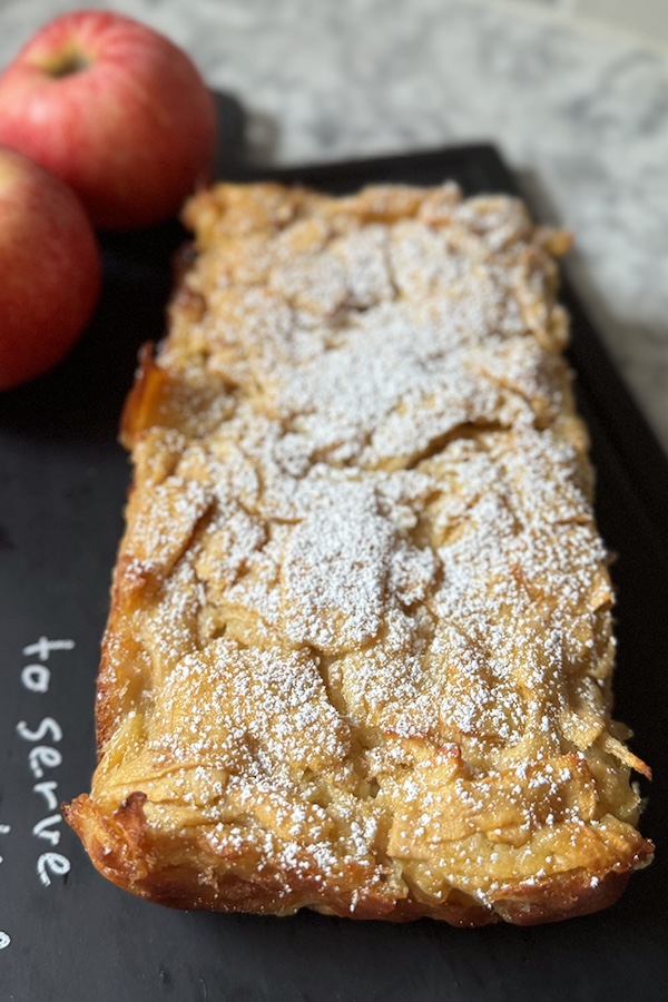 Baked apple pie dusted with powdered sugar on a serving board with whole apples in the background.