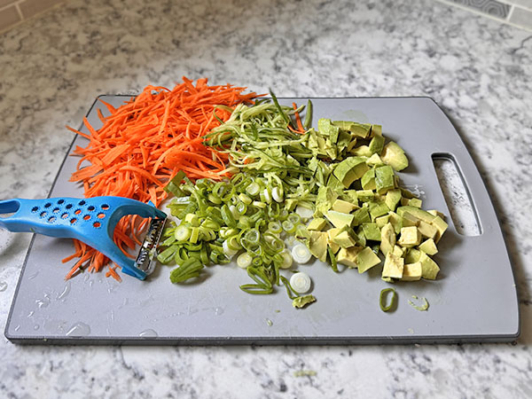 Prepared fresh vegetables: julienned carrot and cucumber, chopped green onion and chopped avocado on a cutting board.