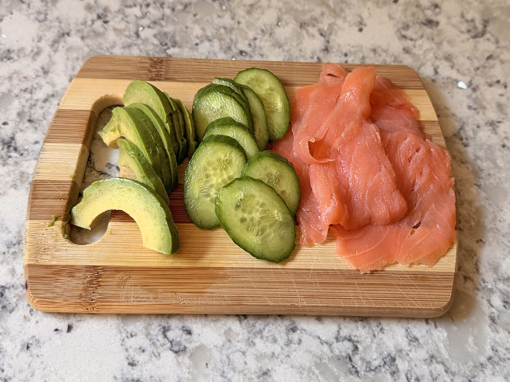 Sushi sandwich fillings arranged on a board: sliced cucumber, avocado, and smoked salmon.