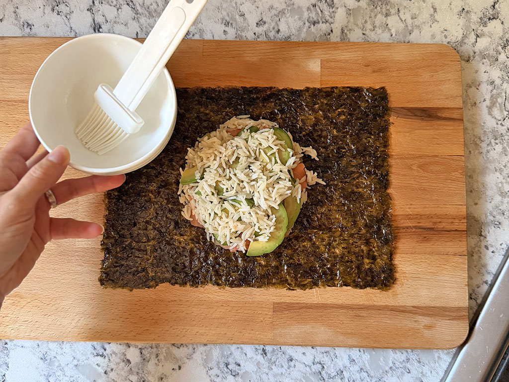 Using a brush to lightly moisten the edges of nori with water before sealing sushi.