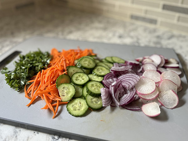 Julienned carrots, sliced cucumbers, radishes, red onion and chopped cilantro on a cutting board.