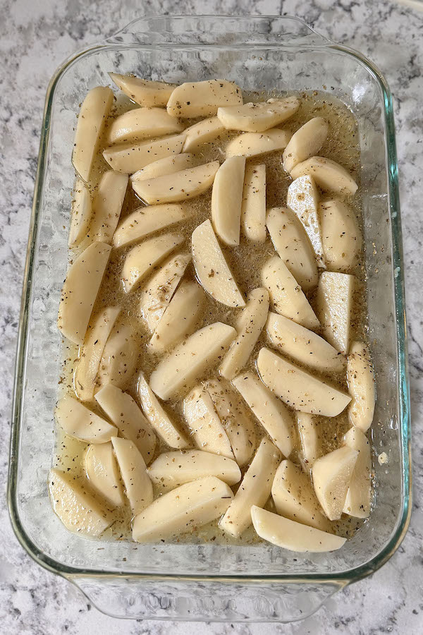 Raw potato wedges in a baking dish, seasoned with chicken broth, lemon juice, oil, garlic, oregano, salt, and pepper.