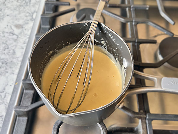 Creamy custard with added orange juice with a whisk in a saucepan on the stovetop.