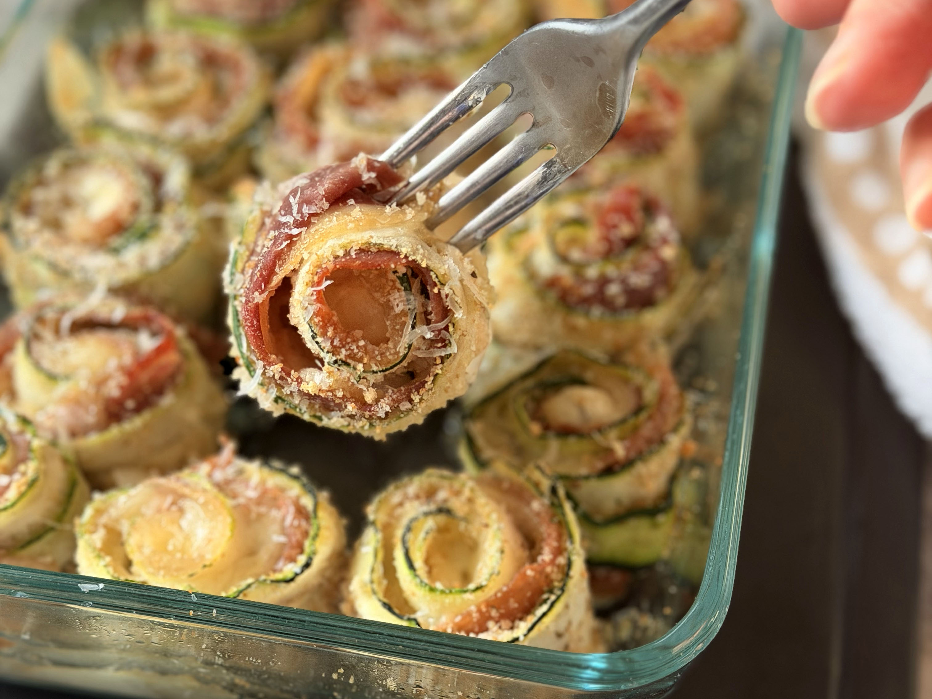 Golden zucchini roll-up held with a fork, with melted cheese and prosciutto inside.