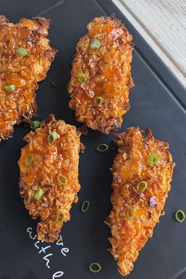 Oven fried cornflake crusted chicken with spicy honey on a serving board, topped with chopped green onions.