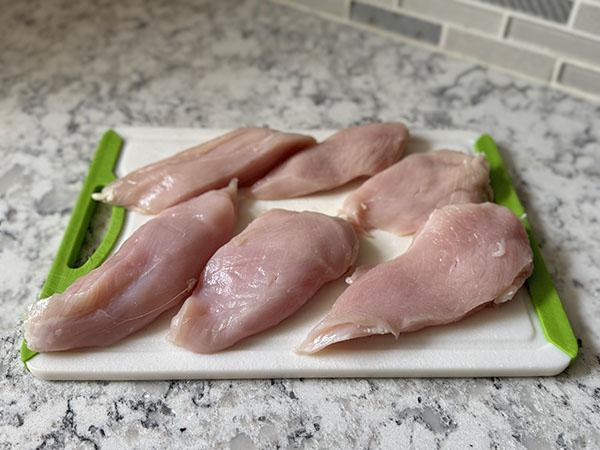 Two chicken breasts, each cut into 3 even-sized pieces on a cutting board.