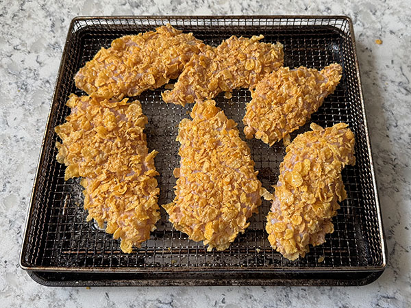 Corn flakes coated chicken pieces on a baking sheet with a wire rack, ready for baking.