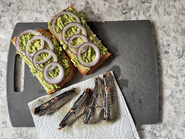 Bread slices topped with mashed avocado and red onions, with sardines draining on paper towels nearby.