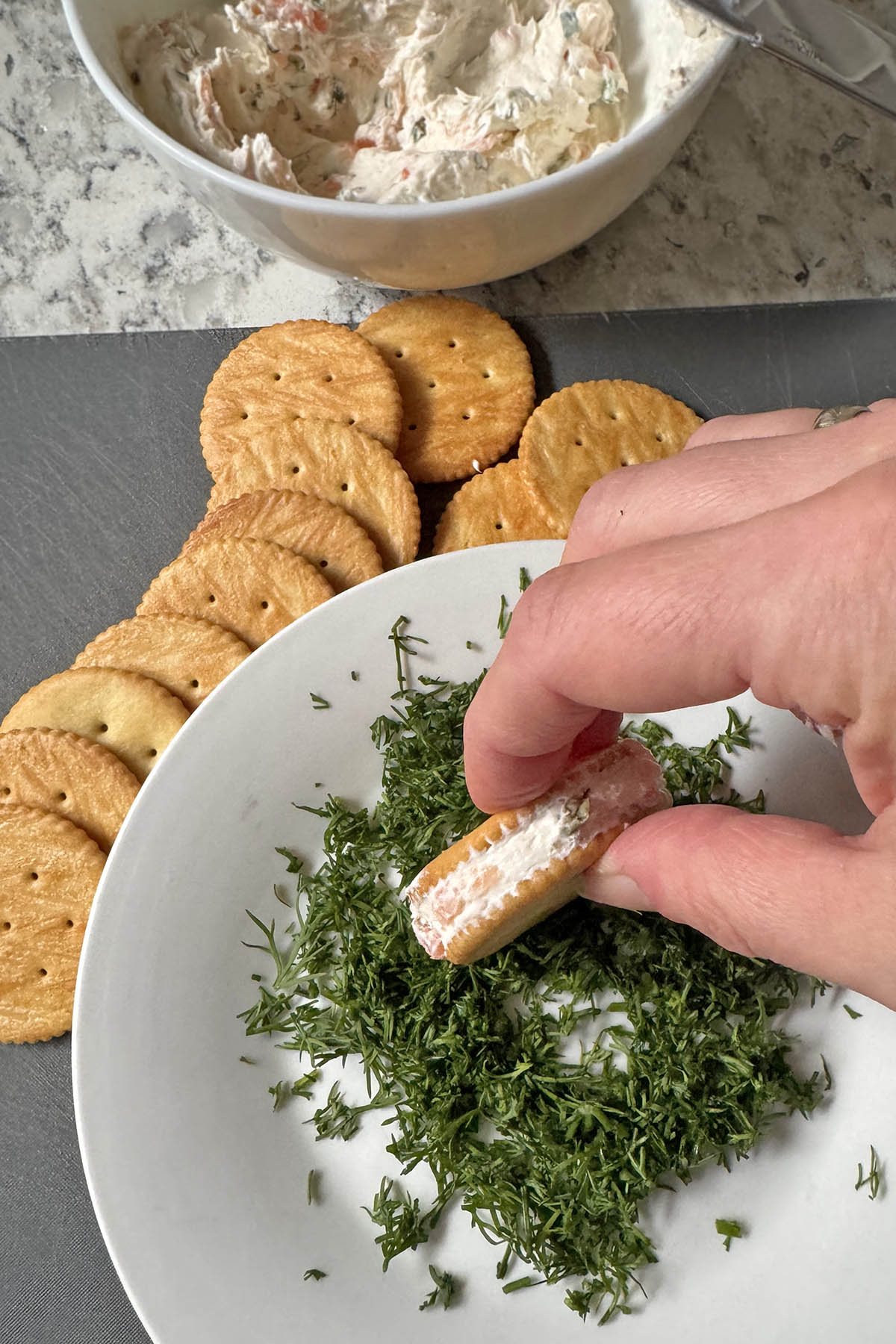 One cracker spread with salmon and cream cheese dip, being rolled in finely chopped fresh dill, with more crackers sitting next to the plate.