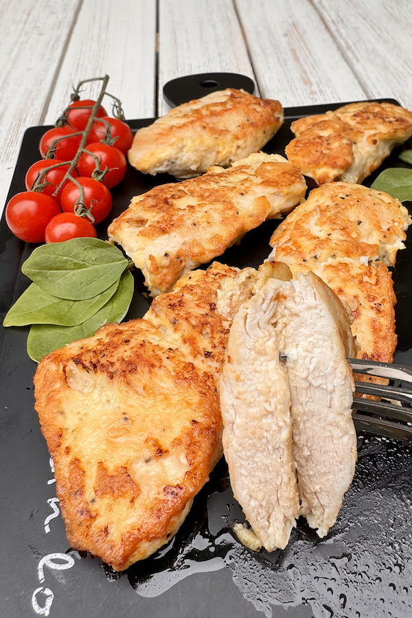 Tender sautéed chicken cutlets on a serving board along with spinach and tomatoes, one cutlet cut in half to show the juicy inside.